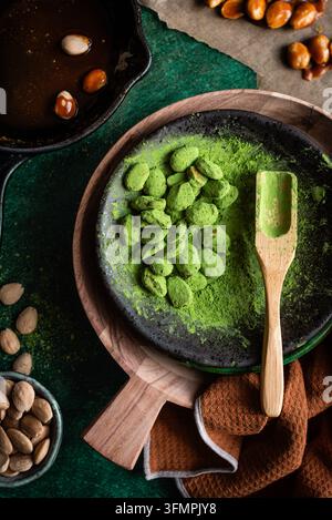 Nature morte vue de dessus avec une poudre de thé matcha vive, des amandes confites vertes, une cuillère en bois et des amandes entières disposées sur des assiettes en céramique sombre texturées Banque D'Images
