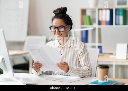 Jeune femme métisse heureuse travaillant au bureau à domicile, examinant des documents, souriante Banque D'Images