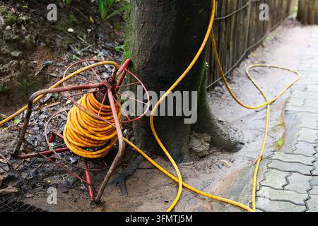Un câble d'extension orange enroulé à côté d'un tronc d'arbre sur un chemin de jardin boueux avec une clôture rustique et du pavage, créant une scène de maintenance extérieure enchevêtrée avec v Banque D'Images