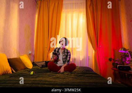 Fille en chemise à carreaux profitant de la musique grâce à des écouteurs sans fil et dansant légèrement sur le lit. Ambiance de coucher de soleil dans la pièce et expression émotionnelle. Vue en contre-plongée Banque D'Images