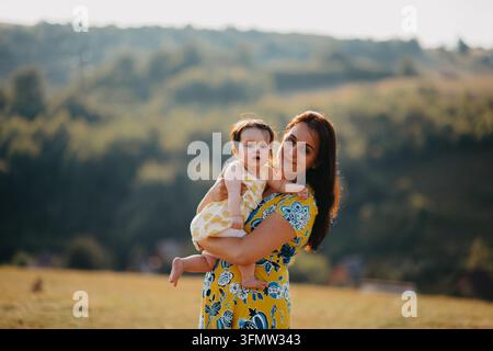 Mère tenant un bébé en robe jaune en plein air par un après-midi ensoleillé à la campagne Banque D'Images