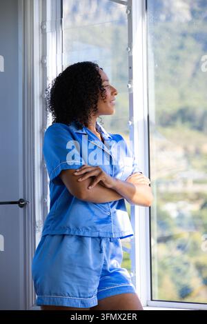 Femme afro-américaine debout, les bras croisés, en pyjama bleu, regardant par la fenêtre avec stores Banque D'Images