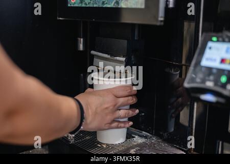 Main d'une personne tenant une tasse en papier blanc alors que le café chaud s'écoule d'un distributeur automatique en libre-service, capturant la routine matinale rapide, la commodité Banque D'Images