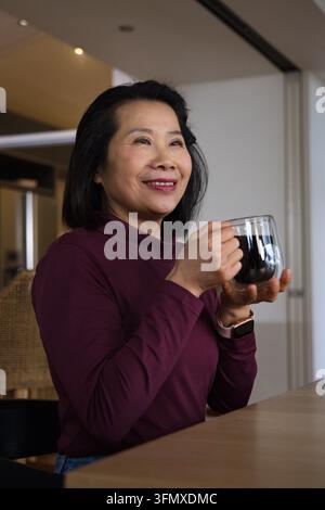 Femme asiatique d'âge moyen tenant une tasse à double paroi à la table de la cuisine, portant un haut prune et une montre intelligente Banque D'Images