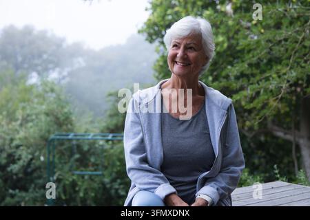 Femme âgée assise sur une terrasse en bois dans le jardin près d'une balançoire verte, portant un sweat à capuche gris et une montre-bracelet Banque D'Images