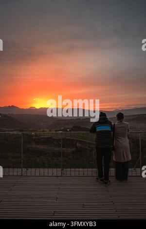 Ronda, Espagne - 11 avr. 2025 : Silhouettés de dos, deux personnes s'appuient à une rambarde sur une plateforme d'observation en bois et contemplent un coucher de soleil rougeoyantsur les montagnes Banque D'Images