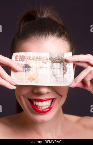 Une jeune femme souriante tient une banque britannique de dix livres devant ses yeux dans un portrait studio ludique, suggérant la richesse, le consumérisme, la confiance financière Banque D'Images