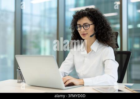 Jeune femme portant un casque et des lunettes, fournissant un service client en ligne et un support technique tout en tapant sur un ordinateur portable dans un environnement de bureau moderne Banque D'Images