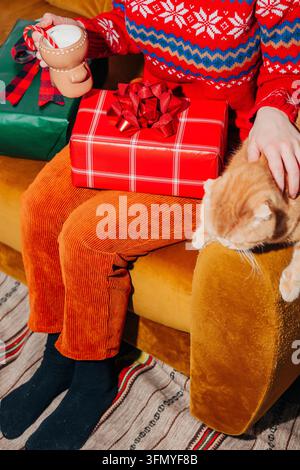 Scène de vacances chaleureuse d'une femme dans un pull festif et un pantalon en velours côtelé jaune tenant du lait à côté d'un chat gingembre, avec un cadeau de Noël emballé sur un b Banque D'Images