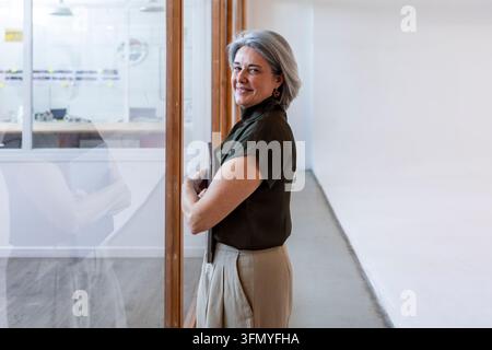 Femme d'affaires mûre et confiante se tient à côté d'une porte de bureau en verre dans un lieu de travail moderne, souriant à la caméra avec un style professionnel informel et Banque D'Images
