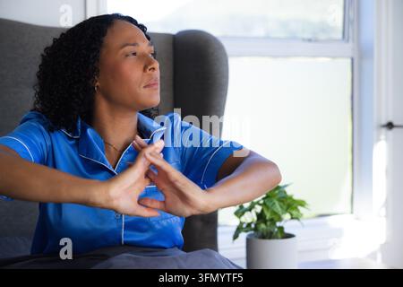 Femme africaine assise dans une chambre, portant un haut de pyjama en satin bleu, tendant les mains vers une plante en pot Banque D'Images