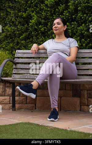 Femme assise sur un banc en lattes de bois dans un jardin aménagé portant une chemise gris clair Banque D'Images