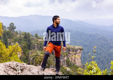 Un randonneur vêtu d'un pantalon orange et d'un haut bleu marine se tient sur une falaise rocheuse en montagne, contemplant une vallée forestière brumeuse lors d'une aventure en plein air. Banque D'Images