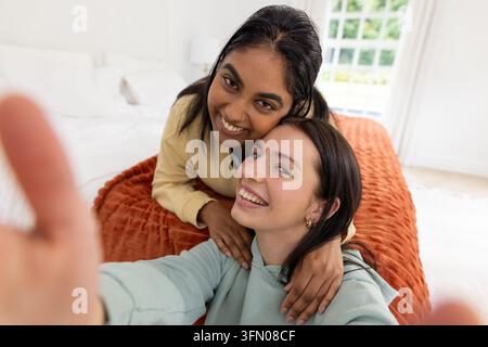 Deux amies posant et souriant en prenant un selfie sur un lit avec une couverture en laine rouille et un smartphone Banque D'Images