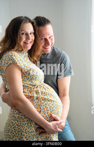 Une mère enceinte et son partenaire se détendent ensemble, s'embrassant dans une pièce calme avec une connexion paisible et sereine à l'intérieur. La mère et le père enceints profitent d'un moment calme et plein d'amour près de son ventre sous une lumière douce. Banque D'Images