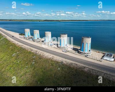 Vue aérienne montrant cinq tours de refroidissement en béton près d'un grand réservoir. Les tours s'élèvent haut, entourées d'une eau bleue calme et d'une terre herbeuse verte. Route ru Banque D'Images