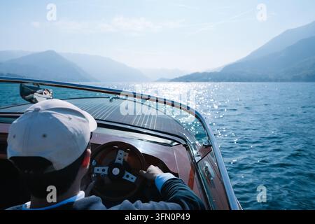 Lac de Côme, Italie – avril, 05, 2026 : Homme avec casquette blanche dirigeant un bateau à moteur en bois sur le lac pittoresque de Côme Banque D'Images