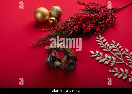 Cliché en studio mettant en scène des figurines de crèche miniature, des boules dorées et des brins de baies rouges disposés sur un fond rouge vif, parfait pour les fêtes Banque D'Images