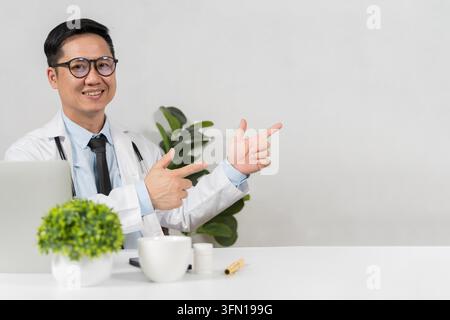 Jeune médecin asiatique sourit en pointant un espace vide. Il porte des lunettes et une blouse blanche. Une plante verte est posée à côté de lui sur le bureau. Idéal pour les soins de santé, m Banque D'Images