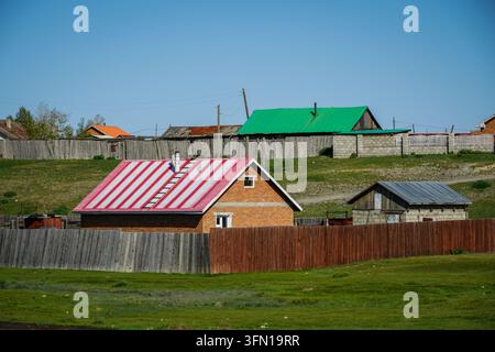 Une maison en briques avec un toit métallique rayé rouge et blanc et une cheminée grise se dresse derrière une clôture en bois brun au premier plan. Derrière elle, une méta verte Banque D'Images