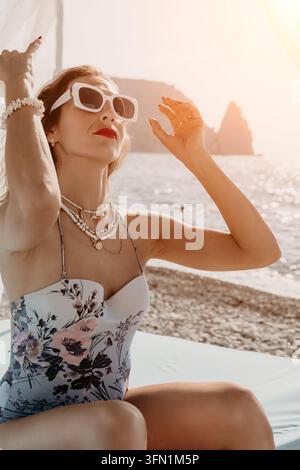 Femme plage coucher de soleil personne élégante en maillot de bain floral et lunettes de soleil blanches assise sur un lit de plage pendant les vacances d'été Banque D'Images