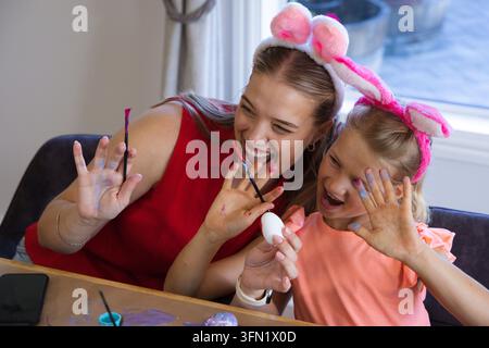 Mère et fille peignant un œuf blanc à la table de la salle à manger à la maison, portant des oreilles de lapin roses et tenant un pinceau Banque D'Images