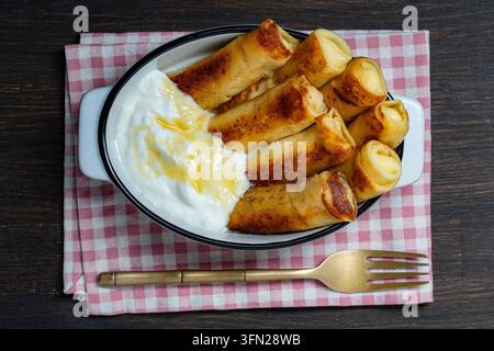Délicieuses crêpes frites farcies au fromage blanc, servies avec du miel sucré et de la crème fraîche dans un plat en céramique blanche sur une serviette à carreaux rustique. Perfect mo Banque D'Images