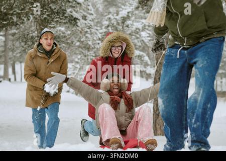 Des amis divers s'amusant à faire de la luge dans une forêt enneigée en hiver Banque D'Images