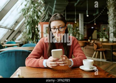 Femme avec lunettes utilisant un smartphone tout en buvant du café dans un café Banque D'Images