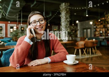 Femme heureuse parlant au smartphone assise dans un café avec un café Banque D'Images