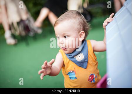 Un enfant joyeux joue dans un espace extérieur vibrant, vêtu d'un débardeur coloré et d'une écharpe mignonne. L'atmosphère animée est renforcée par un adulte à proximité Banque D'Images