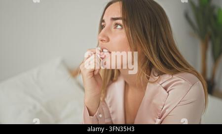 Jeune femme sur un lit mordant ses ongles avec les doigts dans un studio, portant un haut de pyjama en satin et une expression inquiète ; anxiété. Banque D'Images