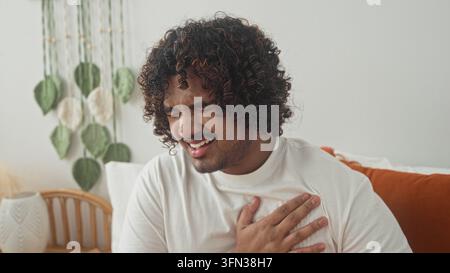 Jeune homme indien serrant la poitrine et appuyant main contre poitrine sur le lit dans le studio avec table de chevet et smartphone, suggérant heartattack ; douleur. Banque D'Images
