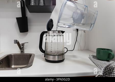 Une main verse de l'eau filtrée d'une cruche dans une bouilloire électrique sur un plan de travail de cuisine. La qualité de l'eau potable et la purification domestique soutiennent la sécurité Banque D'Images