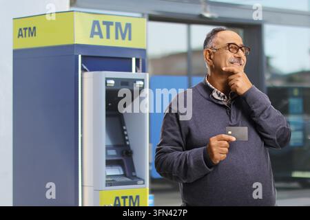 Homme âgé pensif tenant une carte de crédit à côté d'un distributeur de billets Banque D'Images