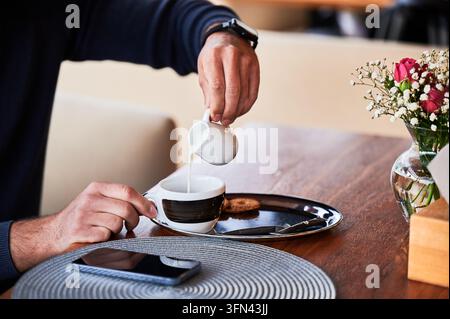 Homme versant du lait dans une tasse de café à une table de café, préparant une boisson chaude pendant la pause, gros plan des mains, cadre cosy, concept de routine matinale et de détente Banque D'Images
