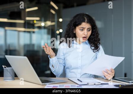 Jeune femme d'affaires à l'expression choquée tenant des documents tout en étant assise à un bureau, réagissant à une mauvaise nouvelle inattendue ou à un problème financier Banque D'Images