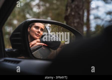 Voyageuse pensive reposant sa tête sur la portière de la voiture, reflétée dans le rétroviseur, au milieu d'un paysage boisé Banque D'Images