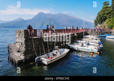 Varenna, Italie – 5 avril 2026 : Les touristes se tiennent le long d'une jetée en pierre surplombant le petit port avec des bateaux amarrés sur le lac de Côme Banque D'Images