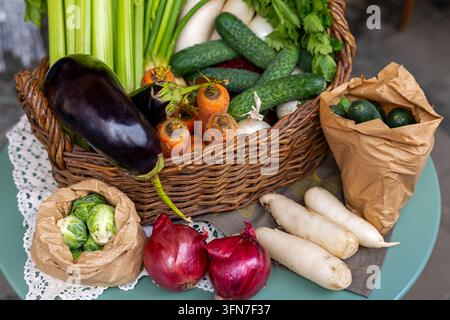 Légumes biologiques frais disposés dans un panier en osier et des sacs en papier sur la table, récolte colorée d'aubergines, de concombres, de carottes, d'oignons et de radis. Banque D'Images