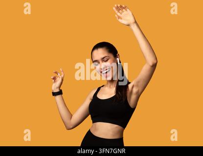 Chanson préférée. Portrait d'une jeune femme sportive excitée portant des écouteurs sans fil écoutant de la musique et dansant isolée sur un fond orange en studio Banque D'Images