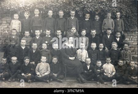Portrait de groupe d'environ quarante écoliers disposés en quatre rangées avec leur instituteur assis au centre, devant un haut mur de briques envahi par du lierre grimpant, à Bečej, en Vojvodina, vers 1920, Royaume des Serbes, Croates et Slovènes. L'inscription au verso indique "Brankovics Mariska gyermekeivel" (Hongrois : "Mariska Brankovics avec ses enfants"), identifiant la femme assise ; les enfants sont probablement ses élèves ou ses pupilles. Mode enfantine des années 1920 : cols marins, pantalons aux genoux, manteaux boutonnés. Tirage vintage original avec les traces d'usure dues au temps, provenant d'un archive familial privé. Banque D'Images