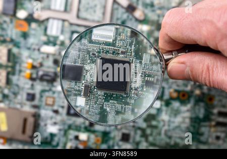 Main d'adulte tenant une loupe inspectant une puce électronique sur un circuit imprimé, illustrant le test électronique, l'analyse matérielle et la technologie m Banque D'Images