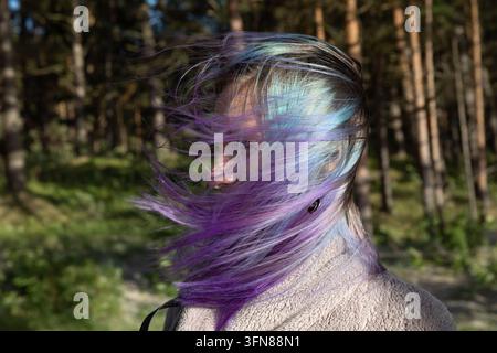 Une femme ressentant le vent qui souffle sur ses cheveux colorés avec des mèches violettes et bleues en plein air parmi les pins. Banque D'Images