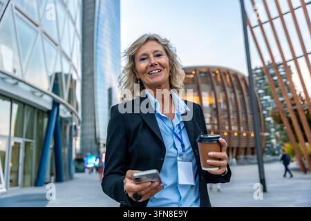 Femme d'affaires professionnelle souriant avec assurance, tenant une tasse de café et un smartphone tout en se tenant dans un quartier d'affaires urbain moderne de Milan, Ital Banque D'Images