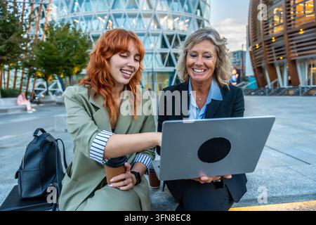 Deux femmes d'affaires joyeuses collaborent sur un ordinateur portable, échangent des idées et discutent d'informations lors d'une réunion informelle en plein air dans une ville moderne Banque D'Images