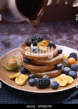 Du sirop d'érable est versé sur une pile de crêpes maison garnies de figues fraîches, de myrtilles, de mûres et de baies dorées Banque D'Images