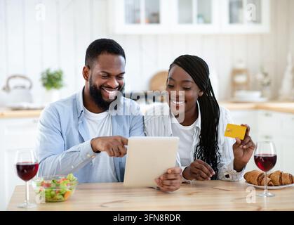 Commander des aliments en ligne. Heureux couple noir avec tablette numérique et carte de crédit dans la cuisine achat livraison de repas ou épicerie de shopping à partir d'Internet, Banque D'Images
