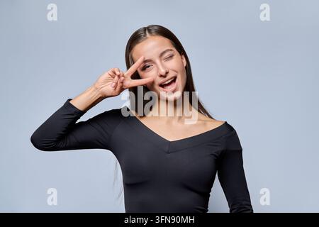 Jeune femme espiègle montrant un signe de paix et clignant de l'œil sur fond de studio Banque D'Images