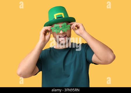 Célébration de la Saint-Patrick, homme joyeux portant un chapeau de leprechaun vert et des lunettes trèfle pour une fête festive sur fond jaune Banque D'Images